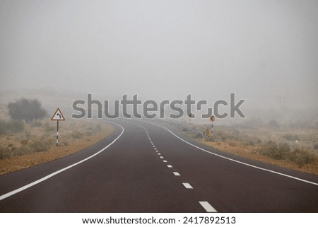 A breath taking landscape view of a curvy road turning left and disappearing in the fog. Left turn sign boards indicating drivers that left turn is ahead be cautious.