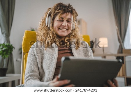 One caucasian woman with curly hair sit at home use digital tablet and headphones to watch movie or series online stream or to have video call happy smile have a good time copy space