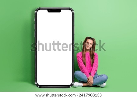 Full size photo of satisfied girl wear pullover sit on floor look at smartphone display empty space isolated on green color background