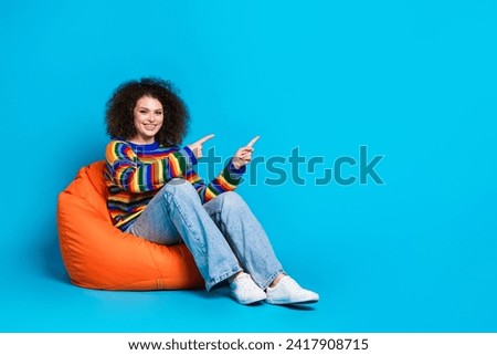 Full length photo of lovely young lady sit bean bag point empty space wear trendy rainbow print garment isolated on blue color background