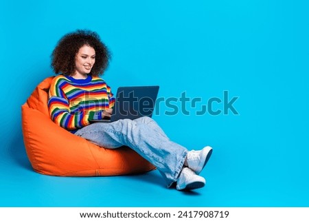 Full length photo of lovely young lady sit beanbag hold netbook wear trendy rainbow print garment isolated on blue color background