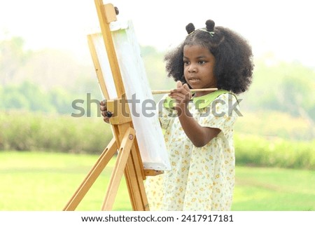 Cute smiling African girl with black curly hair painting on canvas at green garden outdoor. Kid drawing nature picture in green summer park. Happy little child having fun while spending time outdoors.