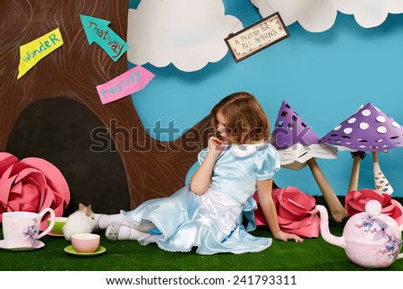 Little beautiful girl with long wavy hair in a blue silk dress in the scenery of Alice in Wonderland with white fluffy bunny cartoon fairy tale adventure