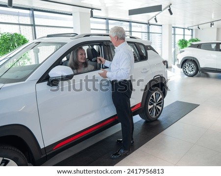 Mature Caucasian woman sits in a new car, an elderly man gives her the keys. 