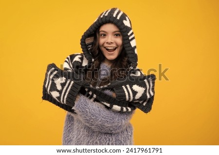Warm hat with hood and scarf. Amazed teenager. Beautiful teen girl in a winter hat and a warm sweater. A child on a yellow isolated background. Excited teen girl.