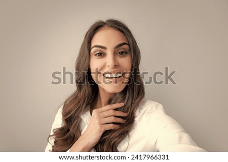 Pleasant girl making selfie in studio and laughing. Good-looking young woman with brown wavy hair taking picture of herself on gray background.