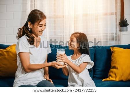 A joyful scene as an Asian mother gives her daughter a glass of milk on the living room sofa. This image radiates affection innocence and the happiness of family breakfasts. Royalty-Free Stock Photo #2417992253