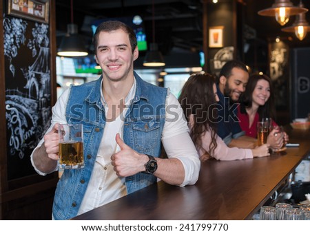Spending time with beer. Portrait of young handsome man who is standing in a pub with glass of beer and smiling. His friends are standing on the background
