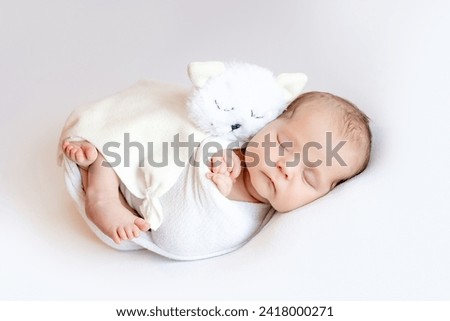A newborn baby girl in a diaper cocoon is sleeping sweetly on a white isolated background with a soft kitten toy. Royalty-Free Stock Photo #2418000271