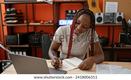Smiling african american woman musician masterfully taking notes on laptop during exciting video call in vibrant music studio