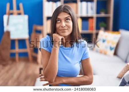 Brunette young woman sitting on the sofa at home with hand on chin thinking about question, pensive expression. smiling with thoughtful face. doubt concept.  Royalty-Free Stock Photo #2418008083
