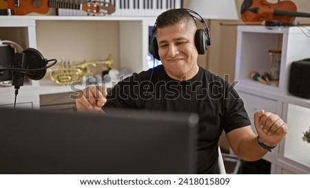 Passionate young latin man, an alluring musician, lost in the melody of his song in the cozy corner of a music studio, dancing while listening with headphones with an infectious smile. Royalty-Free Stock Photo #2418015809