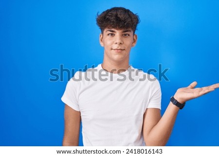 Hispanic teenager standing over blue background smiling cheerful presenting and pointing with palm of hand looking at the camera. 