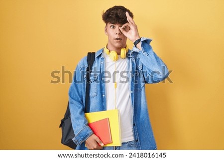 Hispanic teenager wearing student backpack and holding books doing ok gesture shocked with surprised face, eye looking through fingers. unbelieving expression. 