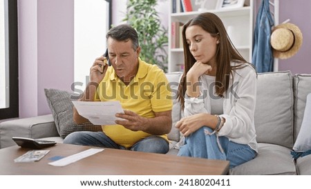 Hispanic father and daughter reading receipt together on their home sofa, serious financial conversation via smartphone Royalty-Free Stock Photo #2418020411