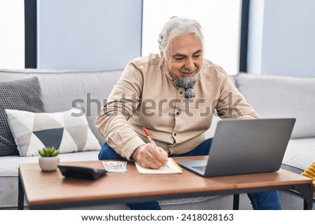 Middle age grey-haired man using laptop writing on notebook at home