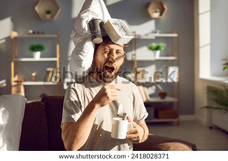 Funny guy can't wake up early in the morning. Young man with a pillow, coffee cup and alarm clock on his head sitting on the couch in the sun light, holding a cup and tea bag, drinking tea and yawning Royalty-Free Stock Photo #2418028371