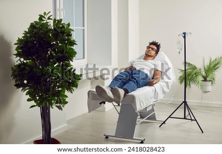 Male patient lying in hospital bed with iv drip attached to his hand. African american male patient receiving IV infusion, getting vitamin treatment in clinic. Medical support and healthcare Royalty-Free Stock Photo #2418028423