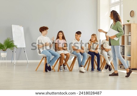 Children psychologist. Friendly young woman conducting psychological training for a group of elementary school children sitting in a row in the classroom and listening their boy classmate. Royalty-Free Stock Photo #2418028487
