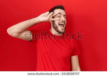 Young hispanic man wearing casual red t shirt doing peace symbol with fingers over face, smiling cheerful showing victory 