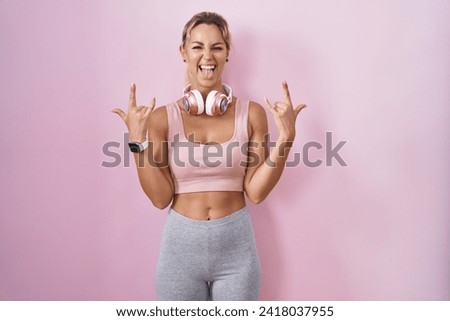 Young blonde woman wearing sportswear and headphones shouting with crazy expression doing rock symbol with hands up. music star. heavy music concept. 