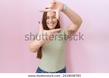 Beautiful brunette woman standing over pink background smiling making frame with hands and fingers with happy face. creativity and photography concept. 