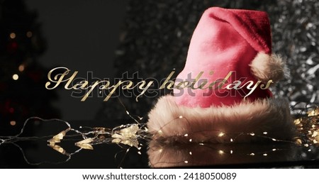 Happy holidays text in gold over christmas hat, lights and tree on dark background. Christmas, tradition, greetings and celebration digitally generated image.