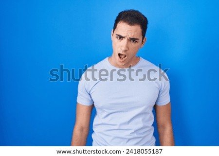 Young hispanic man standing over blue background in shock face, looking skeptical and sarcastic, surprised with open mouth 