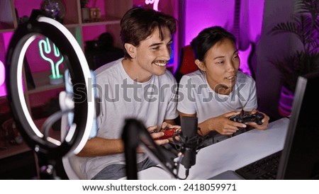 A smiling interracial couple with a man and woman gaming together indoors at night in a room illuminated by purple lights.