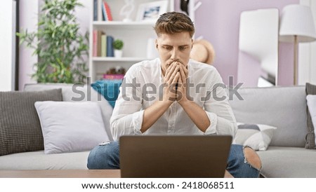 Nervous young caucasian man sitting on a living room sofa, seriously engrossed in using his laptop indoors, deeply focused and worried - a problem in his digital world?