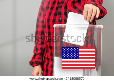 A voter casts a ballot into a ballot box on election day, USA flag. Royalty-Free Stock Photo #2418088571