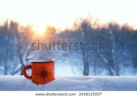 orange enameled traveler tea mug with image of compass wind rose in snow outdoor. winter natural abstract background. symbol of tourism, travel, adventure, hiking. copy space