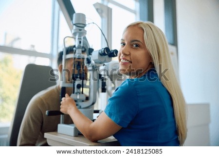 Female ophthalmologist using optical instrument during an appointment with patient at eye clinic and looking at camera. Royalty-Free Stock Photo #2418125085