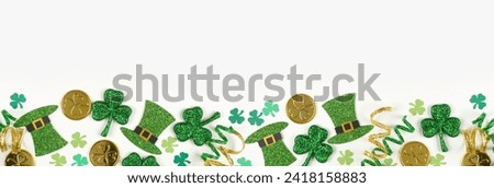 St Patricks Day bottom border of green shamrocks, leprechaun hats, gold coins and ribbon. Top down view over a white banner background with copy space. Royalty-Free Stock Photo #2418158883