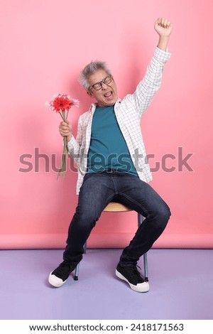 Asian senior man in casual clothing with gesture of siting on the chair while holding a bouquet of flowers isolated on pink background. St Valentine's Day