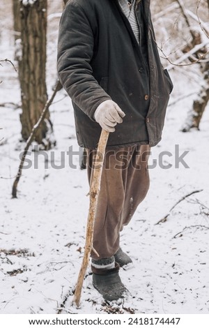 An old, elderly, homeless beggar man, a pensioner in dirty clothes, walks through the forest in nature with a wooden stick in search of food, shelter in the snowy winter in the cold. Poverty concept. Royalty-Free Stock Photo #2418174447