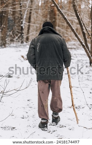 An old, elderly forester, a homeless man in dirty clothes walks through the forest with a wooden stick in search of food, shelter in the cold snowy winter. Royalty-Free Stock Photo #2418174497