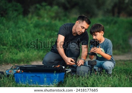 A picture of a caring father showing his son the issues with his electric toy car. The boy is carefully watching what his father is doing while holding the toy's remote control. Outdoor setting, grass