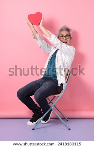 Asian senior man in casual clothing with gesture of siting on the chair while holding holding red heart-shaped isolated on pink background. St Valentine's Day