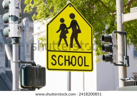 Yellow warning road sign for a school zone at a road intersection with a street traffic light in the background. Yellow School Zone Street Sign, Close-up. School Road Sign with Children, Foreground