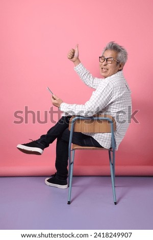 Asian senior man in casual clothing with gesture of siting on the chair while holding mobile phone isolated on pink background. St Valentine's Day