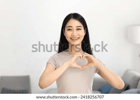 Cheerful attractive young Asian woman showing hand heart, joining finger at chest, promoting love, romance, happiness, friendship, looking at camera for portrait, smiling, laughing