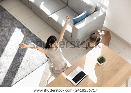 Happy Asian girl enjoying business achievement, education success, celebrating victory at laptop, making yes triumph gesture, sitting at home office workplace table, raising hands up. Top view shot Royalty-Free Stock Photo #2418256717