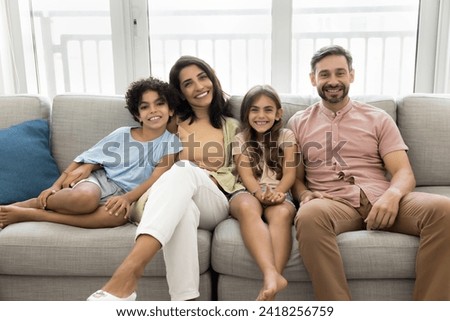 Happy family couple and cheerful sibling kids home portrait. Latin parents and two children sitting close on couch together, looking at camera, smiling, laughing, enjoying leisure in apartment