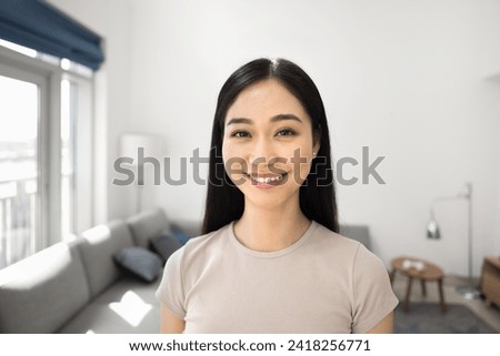Cheerful happy Asian girl home head shot portrait. Young Chinese woman standing in living room, looking at camera with toothy smile, posing in comfortable interior