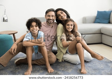 Happy Latin couple of parents and two cheerful kids sitting on carpeted floor at cozy home, looking at camera, posing for family portrait, smiling, laughing, enjoying childhood, parenthood