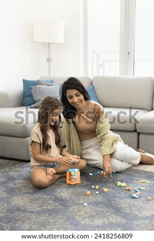 Happy loving young mother and cute daughter child constructing toy tower on home floor, playing family learning game on carpet, stacking plastic building blocks, colorful cubes, smiling. Vertical shot