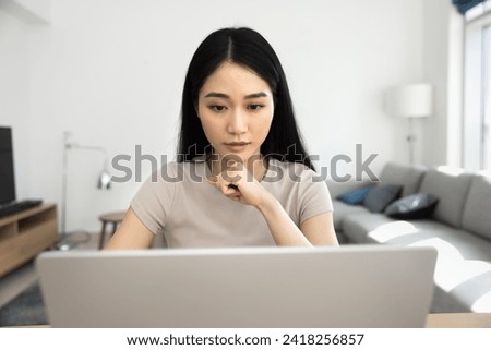 Focused serious young Asian professional girl working on online business project at home, sitting at table with laptop, leaning chin on hand, using Internet technology. Front view Royalty-Free Stock Photo #2418256857