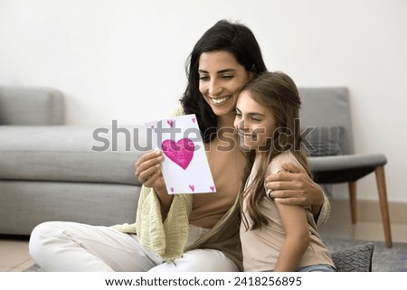Cheerful Latin mom and kid celebrating mothers day at home. Happy mum hugging adorable daughter with love, tenderness, holding paper handmade greeting card with drawn heart, smiling