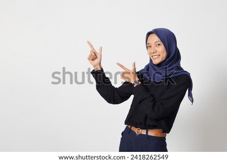 Portrait of excited Asian hijab woman in casual shirt promoting product, pointing finger to the side. Advertising concept. Isolated image on white background